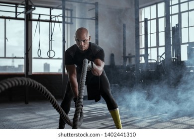 Bald athlete doing battle rope exercise at crossfit gym. Concentrated african man doing cross fit exercise while working out in gym. Focused man in sportswear training exercise.