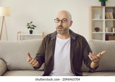 Bald Bearded Adult Man In Glasses Sitting On Sofa, Looking At Camera, Talking And Gesturing. Mature Man Taking Part In Online Meeting Via Video Call. Webcam Portrait, Laptop Computer Screen View