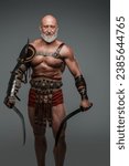 Bald bearded gladiator of mature age is featured in this photo, clad in light armor and wielding two swords while standing before a grey background