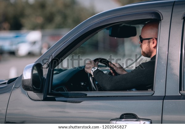 bald and bearded male in glasses with a clock in\
a suit behind the wheel of a\
car