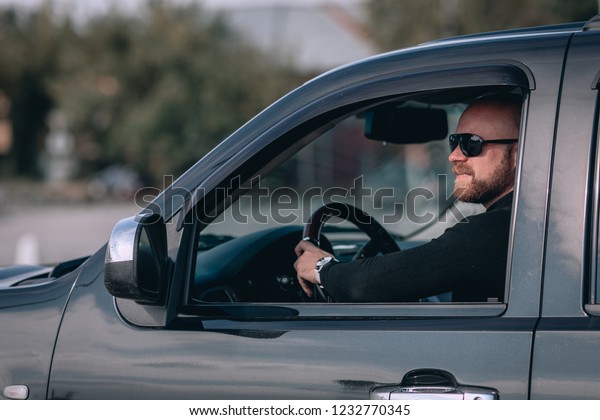  Bald and\
bearded man in glasses with a clock in a suit behind the wheel of a\
 black car. motorists concept