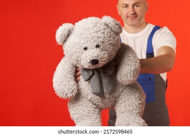 Bald, Bearded Man In A Loader's Suit Carries A Large, Gray Teddy Bear On A Red Background. Dad Is Carrying A Gift For His Daughter On Her Birthday