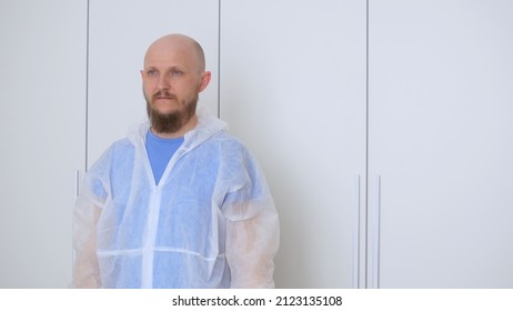 A Bald Bearded Man In A Locker Room In A Protective Suit To Visit The Place Of Origin Of The Coronavirus. The Work Of A Nurse In A Covid Hospital During The Outbreak Of Coronovirus Infection