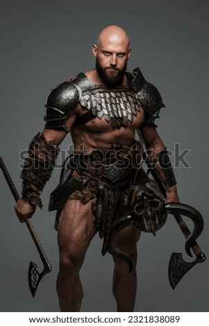 A bald bearded viking in lightweight fur-lined armor holding a two axes stands against a gray background