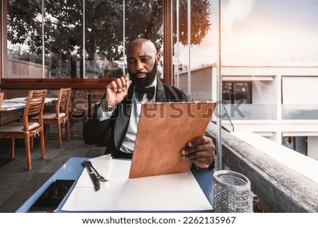 A bald black man dressed in a sleek black tuxedo and bow tie is perusing the cork color menu at a restaurant, located on a rooftop, with a side view while holding a pen; he is looking thoughtful
