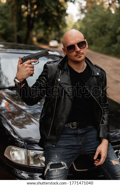 Bald brutal bearded man in sunglasses in black\
fashionable leather jacket stands in front of his car and holds gun\
near his head. Cool bully guy posing near black auto with guns\
outdoors. Bad boy.