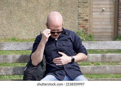 Bald Caucasian man wearing jeans, blue shirt sitting on wooden bench in the park one hand holding sunglasses the other hand on stomach as stomach ace 