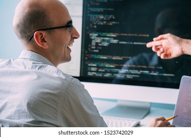 Bald Caucasian Programmer In Glasses Is Sitting Behind The Desk, In Front Of Black Screen, Filled With Code Lines. He's Laughing, Chatting With Female Coworker About Programming.