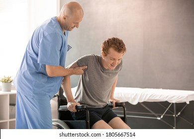 Bald Clinician In Uniform Helping Young Man To Sit In Wheelchair After Rehabilitation Training While Supporting His Arm And Elbow