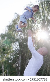 Bald Dad Tosses Up Son In The Field Or Forest On Sunny Summer Day. Father's Day.