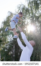 Bald Dad Tosses Up Son In The Field Or Forest On Sunny Summer Day. Father's Day.