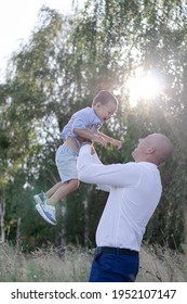 Bald Dad Tosses Up Son In The Field Or Forest On Sunny Summer Day. Father's Day.