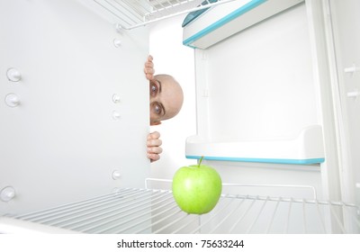 Bald Head Of Young Man Looking Around Corner Of Open Refrigerator Containing Single Green Apple.