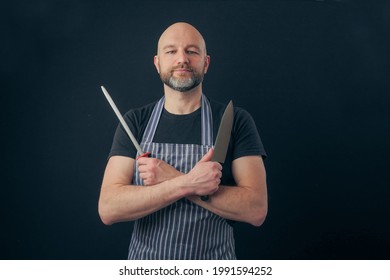 Bald male butcher or fishmonger in black t shirt and classic black and white apron on dark background. Meat industry. Holding knife and metal steel. Man in his 40s, grey beard, smile on his face - Powered by Shutterstock