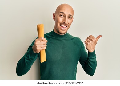 Bald Man With Beard Holding Dry Spaghetti Pointing Thumb Up To The Side Smiling Happy With Open Mouth 