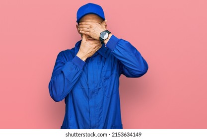 Bald Man With Beard Wearing Builder Jumpsuit Uniform Covering Eyes And Mouth With Hands, Surprised And Shocked. Hiding Emotion 