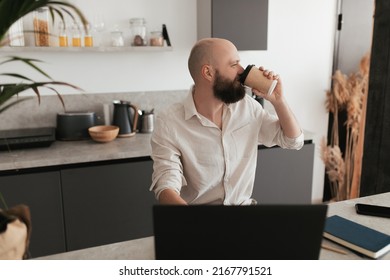 A Bald Man With A Beard Works Remotely In His Modern Kitchen Using A Laptop And Drinks A Hot Drink From A Craft Glass.