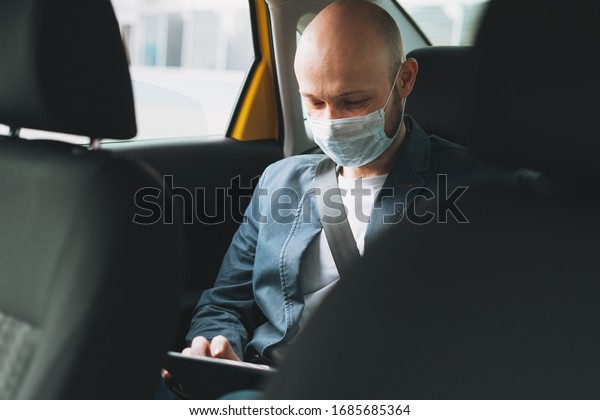 Bald man businessman in medical face mask\
using tablet mobile phone inside yellow car taxi. Concept of\
coronavirus quarantine