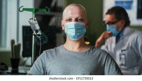 Bald Man With Cancer Wearing Mask And Looking At Camera While Sitting Near Oncologist During Chemotherapy Session In Hospital