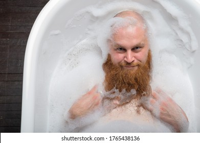 A bald man is fooling around in the foam bath. The guy makes funny hairstyles on his red beard. - Powered by Shutterstock