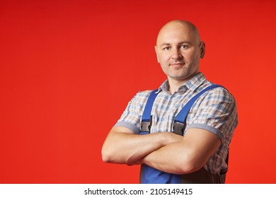 Bald man in overalls and a plaid shirt stands on a red background, confidently crossing his arms over his chest. Portrait of a bald guy looking into the camera.