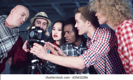 Bald Man Producer With Laptop Talking To Crew Of Young Mixed Ethnic People Filmmakers Standing Near DSLR Camera On Tripod In Dark Empty Hall