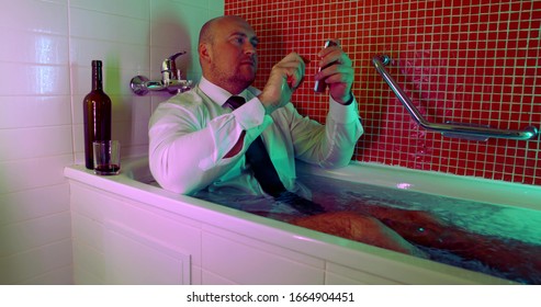 bald man is sitting in bathtub dressed in shirt and tie, typing message on cell phone, drunk and funny - Powered by Shutterstock