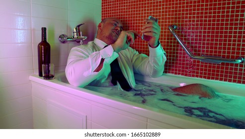 a bald man, sitting in a white shirt and tie in a bathtub filled with water and foam, looking at his mobile phone, smiling, next to him on the edge of the bathroom is a bottle of wine and a glass. - Powered by Shutterstock