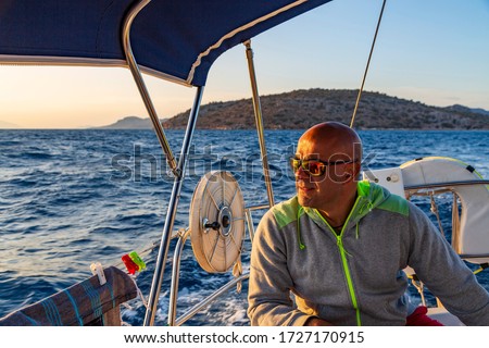 Bald man with sun glasses is sitting on deck of sailing boat and enjoying the summer and his journey. View from stern to blue waters, sky and green island.