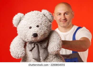 Bald Man In Work Clothes Gives A Large Gray Teddy Bear With A Butterfly On A Red Background. The Courier Brought A Gift, Delivery Of Soft Toys