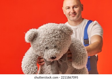 Bald Man In Work Clothes Holds A Gray Teddy Bear On A Red Background. The Courier Brought A Gift, Delivery Of Soft Toys.