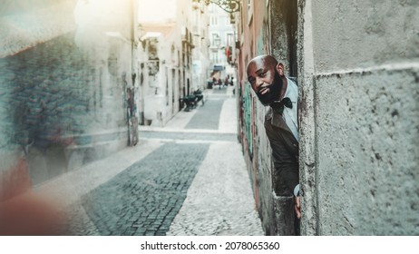 A bald mature bearded African American guy in an elegant suit with a bowtie is leaning out of a doorway on a stoop of an antique street with a paving-stone and a copy space and reflection on the left