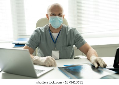 Bald Mature Male Clinician In Protective Mask Looking At You While Sitting By Workplace In Front Of Camera And Using Laptop In Medical Office