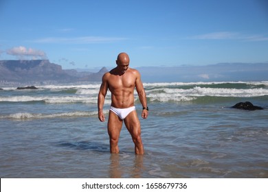 Bald muscled guy in white speedo standing in sea at beach with Table Mountain in background