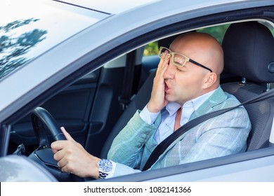 Bald stylish man in glasses rubbing face and looking embarrassed while sitting in car and driving on road having problems with traffic. 
