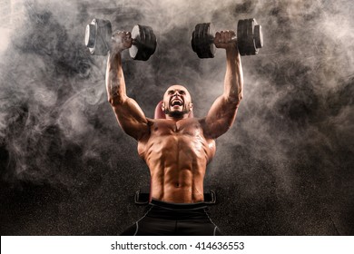 Bald Topless Muscular Man Doing Exercises With Two Dumbbells On Bench Press In Smoke 