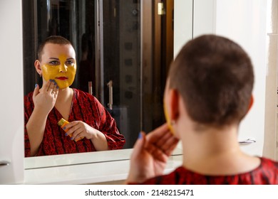 A Bald Woman Applies A Cosmetic Mask On Her Face In The Bathroom. Reflection In The Mirror. Horizontal Photo