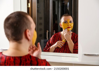 A Bald Woman Applies A Cosmetic Mask On Her Face In The Bathroom. Reflection In The Mirror. Horizontal Photo
