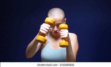 Bald Woman Boxing To The Camera With Dumbbells In Hands On A Dark Background.