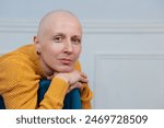 A bald woman cancer survivor with serene expression sitting in a chair, her hand resting on its arm, a white wall in the background