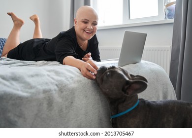 Bald Woman Feeding Her Dog While Laying At The Bed And Working