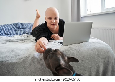 Bald Woman Feeding Her Dog While Laying At The Bed And Working