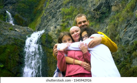A Bald Woman, Her Children And Her Husband Are Hugging On The Background Of A Waterfall, Looking At The Camera.