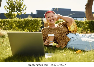 A bald woman relaxes on the grass, smiling while listening to music on headphones with a coffee. - Powered by Shutterstock