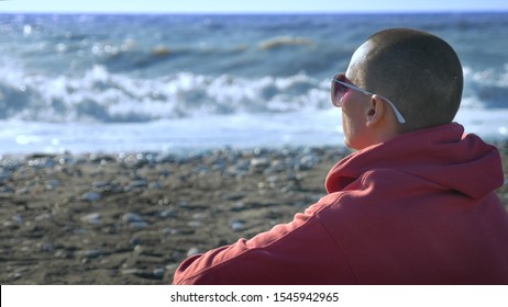 Bald Woman In A Sweatshirt Sits On The Seashore During A Storm.