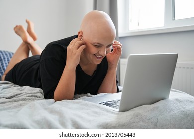 Bald Woman Wearing Earphones Laughing While Laying On The Bed And Working At The Laptop