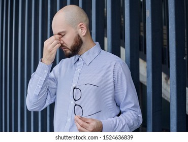 A Bald Young Business Guy With A Brown Beard Stands On The Background Of A Building In The City. Fatigue, Hand On Nose Bridge. Hard Week.