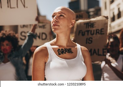 Bald Young Female Activist Protesting Outdoors With Group Of Women In Background. Woman With Word Brave Written On Her Body.