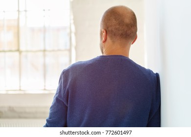Balding man standing staring through a window while leaning on an interior white wall in a close up rear view - Shutterstock ID 772601497