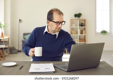 Balding Middle Aged Man Using Laptop. Freelance Worker Or Online Tutor Sitting At Desk In Home Office, Drinking Coffee And Working On Computer. People Browsing Internet, E-learning, Remote Job Concept